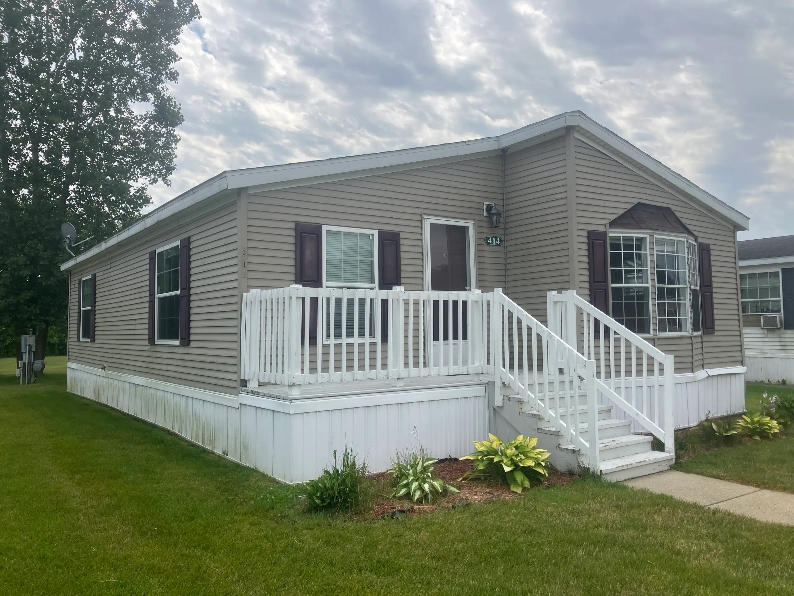 Front view of a beige manufactured home with white railing stairs, dark shutters, and a bay window on a green lawn under a cloudy sky.